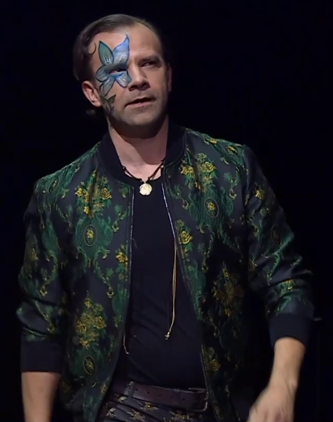 Liam dressed as Orym on a stage, staring curiously off into the distance. He wears a green jacket with ornate gold patterns over a solid black shirt tucked into a brown leather belt. A blue five-petaled flower is painted above his right eye. A necklace with a golden circle hangs just at his clavicle and golden cords dangle down to his stomach.