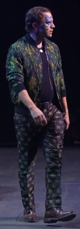 Liam dressed as Orym striding across a stage, staring off into the distance. He wears a green jacket with ornate gold patterns over a solid black shirt tucked into a brown leather belt. A blue five-petaled flower is painted above his right eye. He has dark pants patterned with what appear to be flying insects and brown leather boots. A necklace with a golden circle hangs just at his clavicle and golden cords dangle down to his stomach.