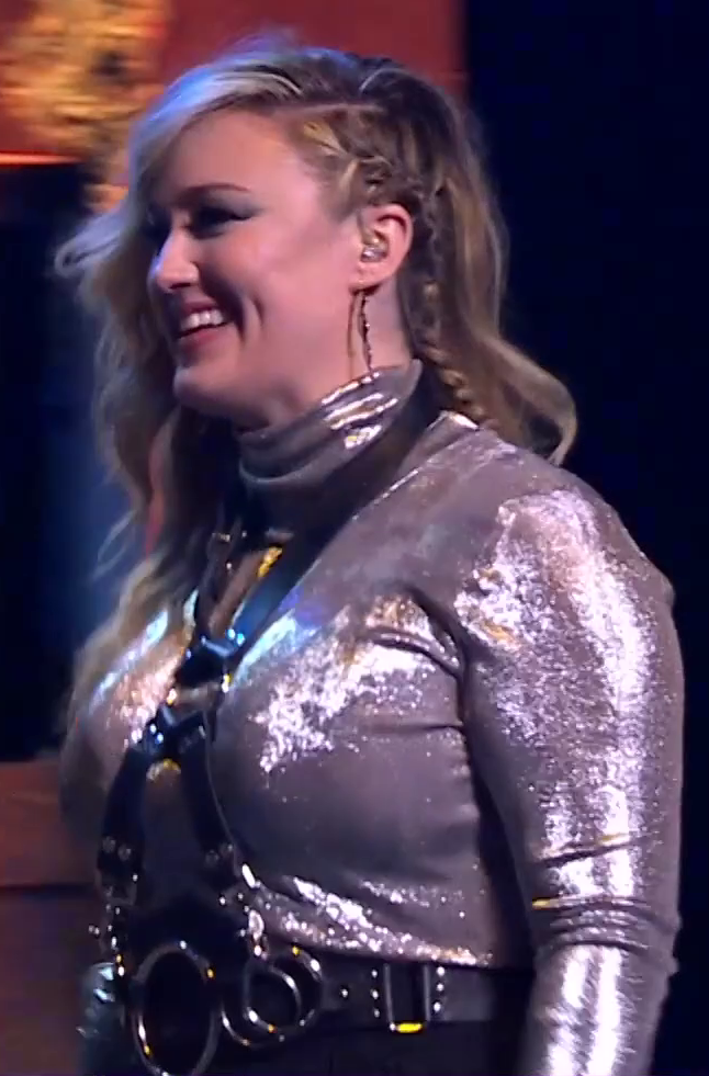 Ashley dressed as Yasha beaming as she walks across a stage. She wears a sparkly silver top with a black leather harness secured on top of it. The harness is joined with large silver rings to a wide black leather belt. A single braid emerges from her otherwise unbraided brown hair. An earpiece is in her left ear.
